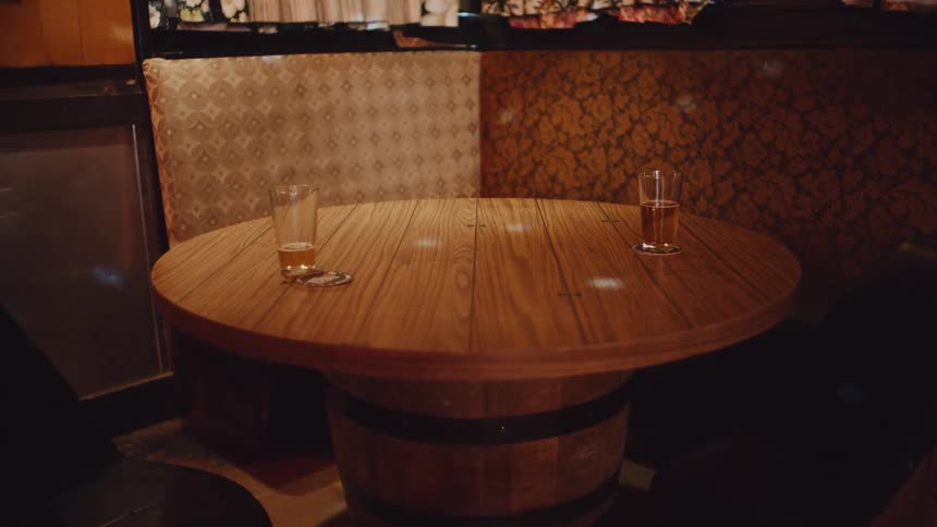 Two drinks sit on wooden table with beer keg base in corner booth near window as dancing lights swirl around