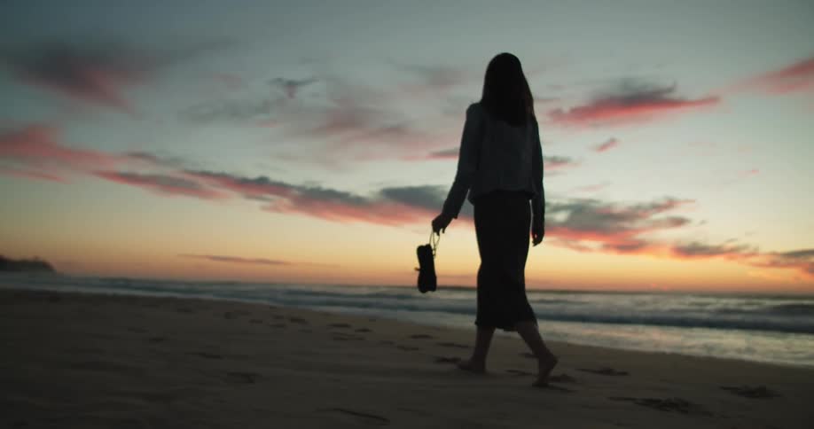 The silhouette of a woman walking on the beach carrying her shoes at sunset