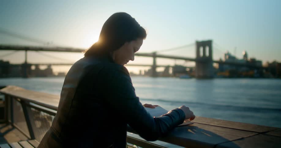 A woman reading a book with a large city bridge in the distance at sunset