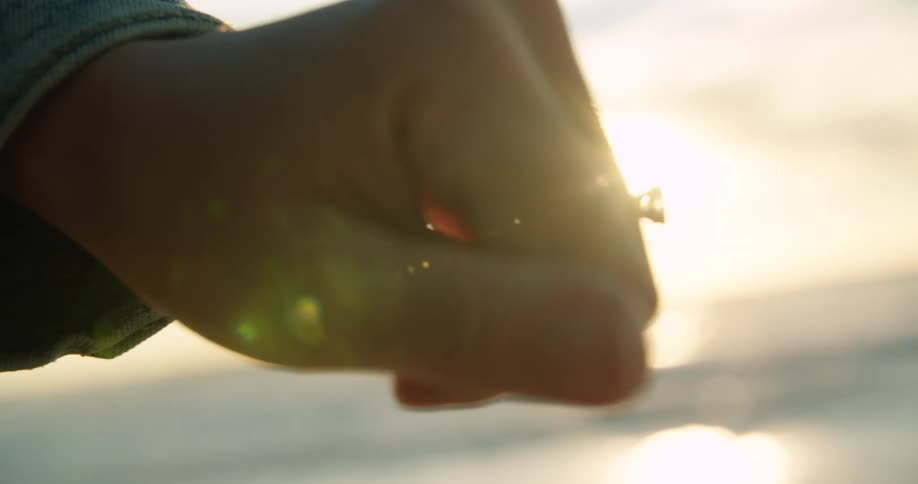 Woman stretches hand out as sun shines from horizon past water and over fingers and diamond ring