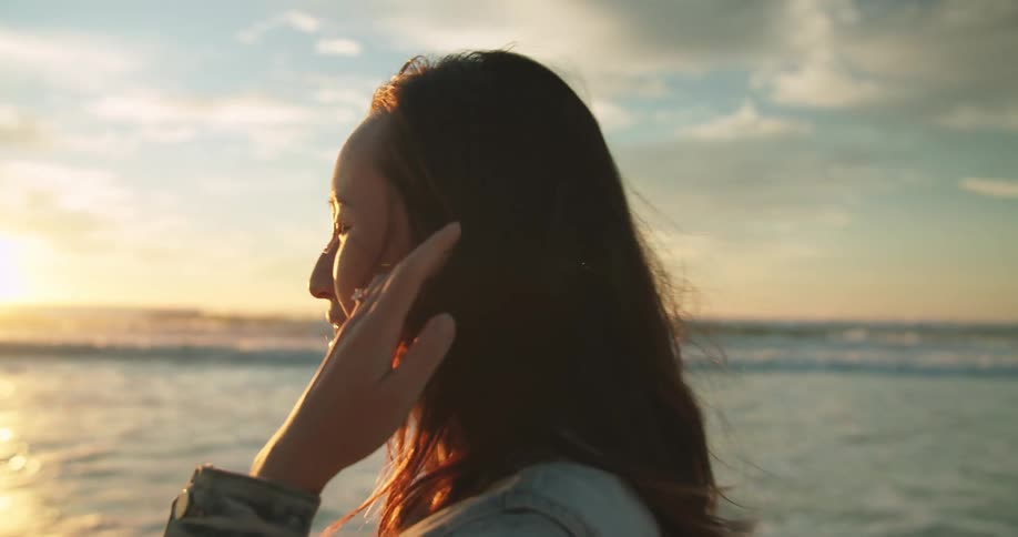 Woman touches her hair while looking at the sunset next to the ocean
