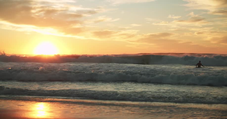 The sun glows on the horizon reflecting off the waves and sand as a man wades into the water with a surfboard