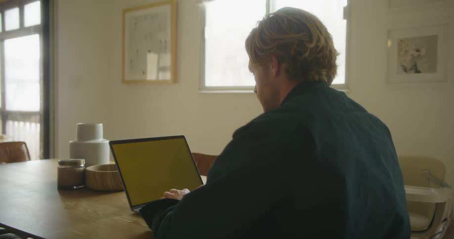 A man sits at his dining room table typing on a laptop