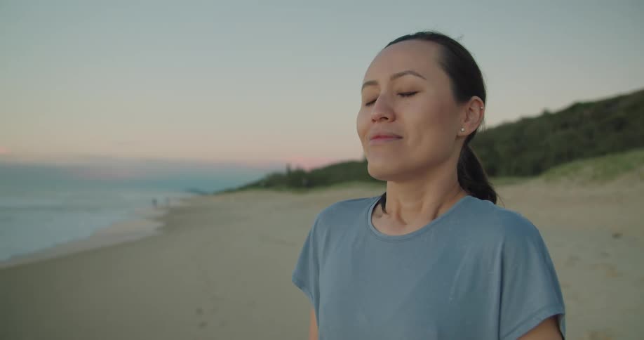 Woman on beach takes deep breath as she smiles with forest covered hill and red tinted horizon in background