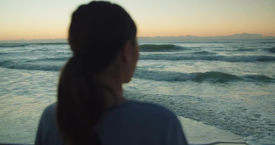 Woman with a ponytail and a t shirt watches the ocean with waves at dusk