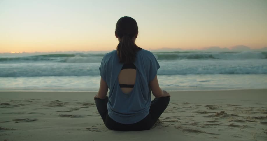 Woman sits cross legged on sand at beach looking toward red sky on the horizon as waves roll onto shore