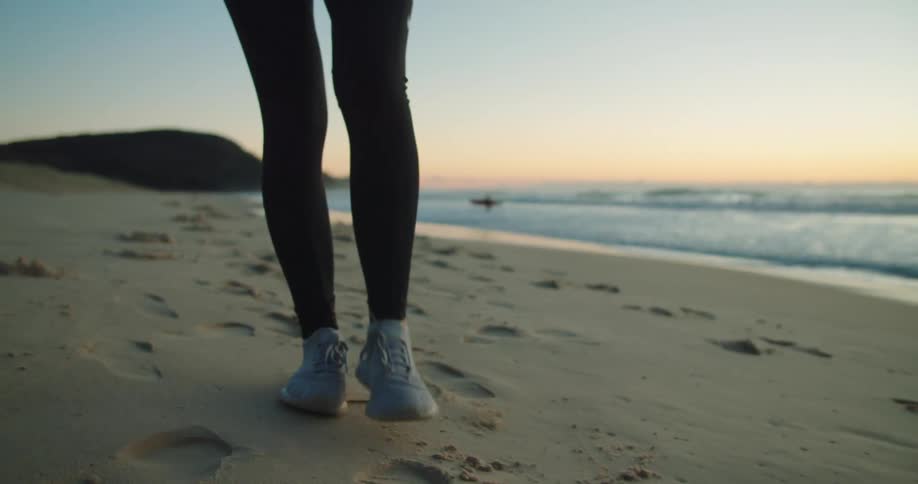 Person wearing tights walks along beach leaving footprints as surfer in distance walks into ocean