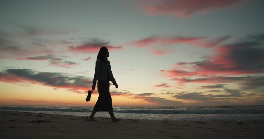Barefoot woman wearing a dress walking on the sand next to the ocean at sunset