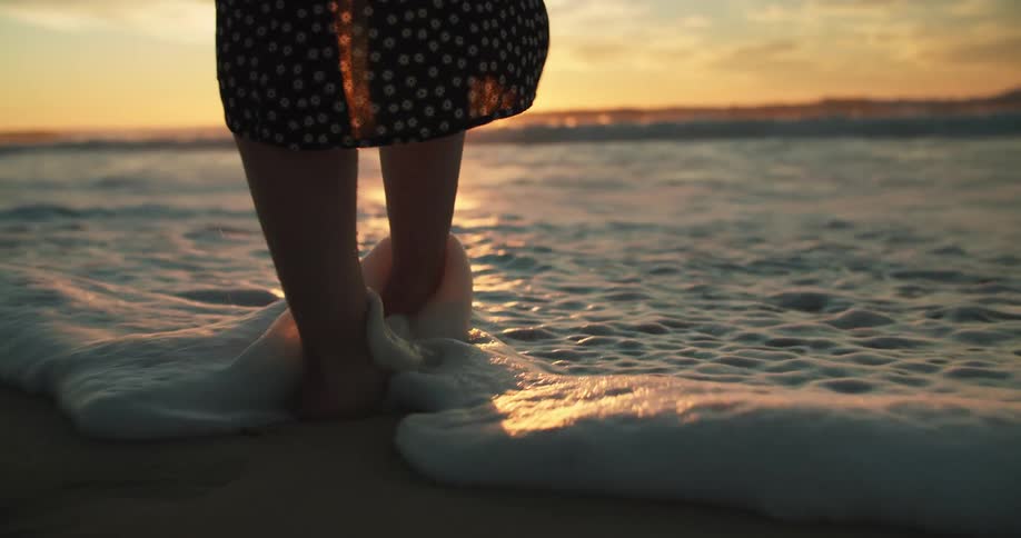 A woman dips her toes in water at the coast of a beach at dusk