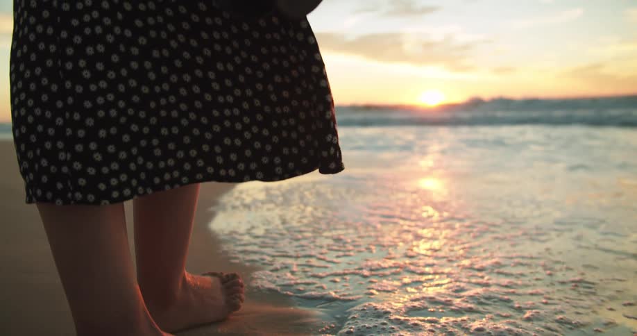 A woman standing barefoot on a sandy beach with ocean waves washing over her toes at sunset