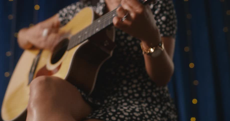 A female guitarist wearing a dress and cowboy boots sits while strumming on her acoustic guitar
