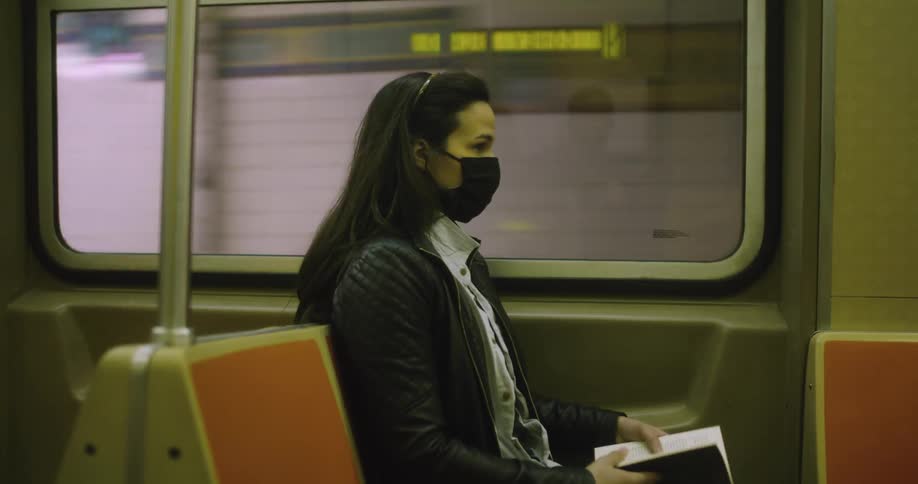A woman in a face mask flipping through a book while riding on a subway train