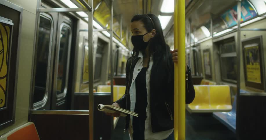 Woman with a black mask and a denim jacket reads a book on the subway train