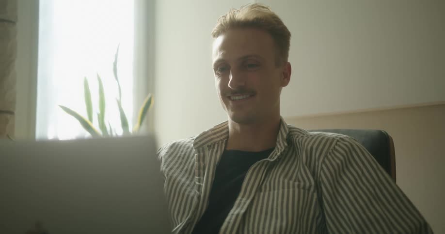 Man with a moustache and a shirt sits at table and makes a video call on grey laptop in a room