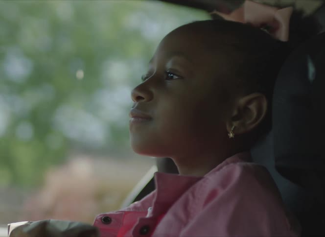 A young girl in a pink jacket and bow rides in the backseat of a vehicle as it drives through a neighborhood