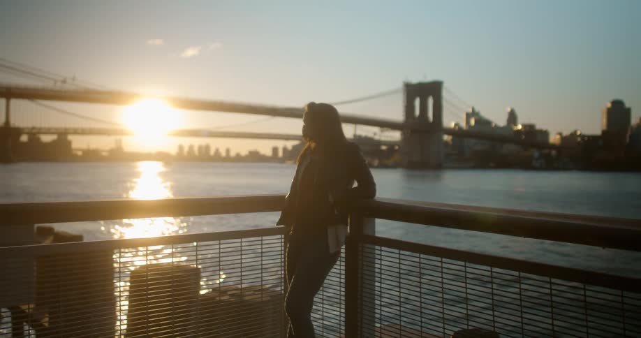 A woman leaning against railing with a city bridge in the distance at sunset