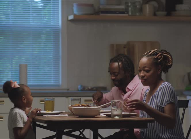 A man a woman and a girl in a room sitting on a table and eating with a wall and window in the background