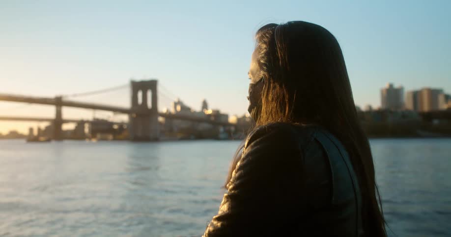 A woman wearing a face mask stands along the side of a river across from a bridge and metropolitan area