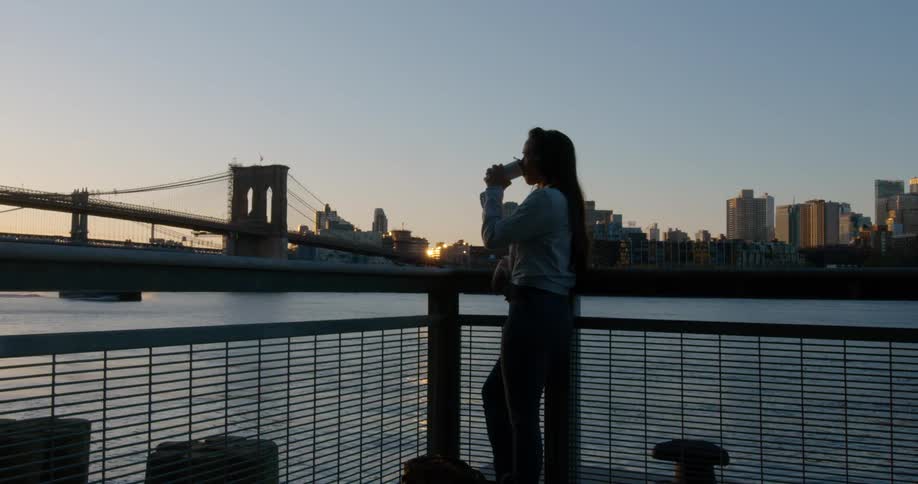 A female standing outside on a deck near the water drinking from a cup