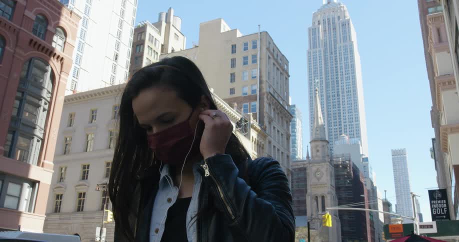A woman in a face mask putting in earbuds outside on a city sidewalk