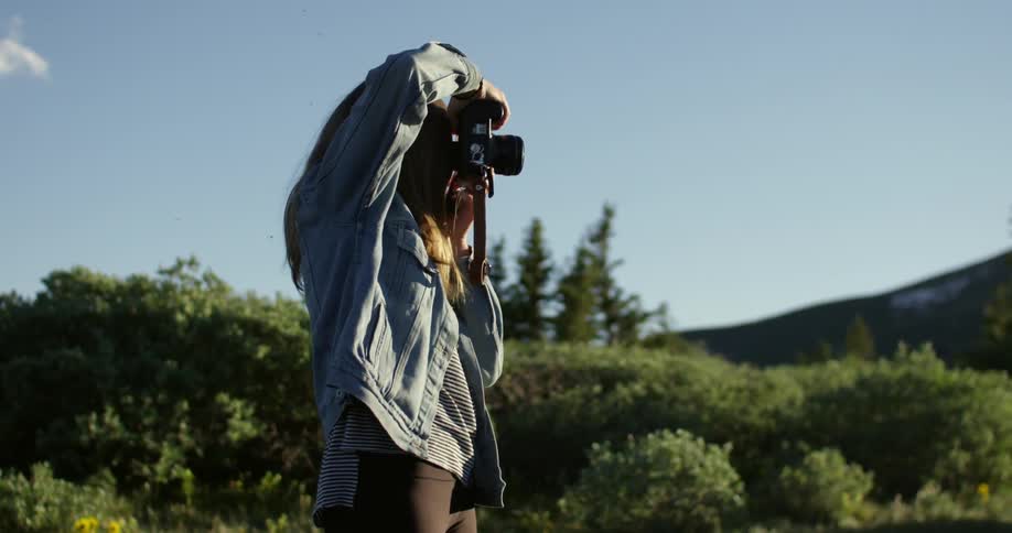 A woman stands in the grass and shrubs poised to take a photograph
