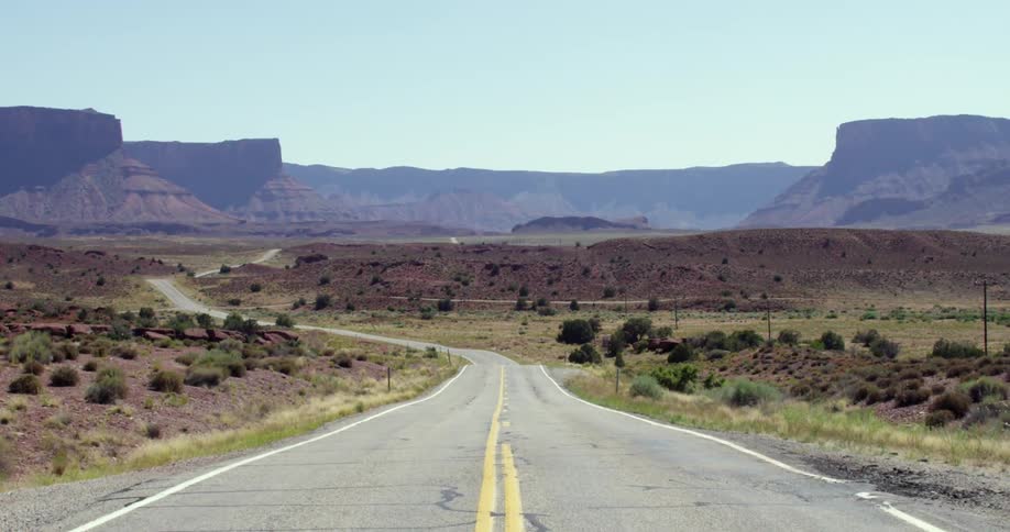 A long paved road in the desert with several mesas in the distance