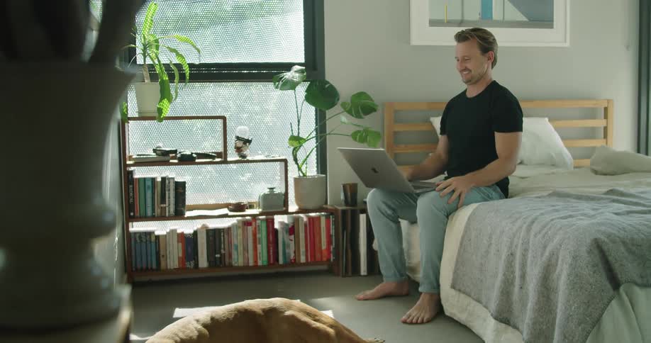 Man sitting on bed makes a video call on his laptop next to a brown dog in the bedroom