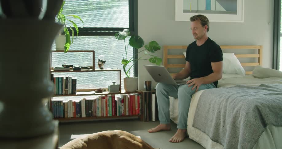 A man is sitting on a bed and typing on a laptop