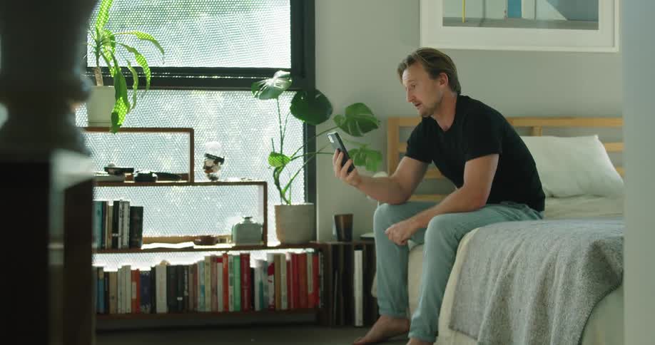 A man sitting on a bed looking at a smart phone with a dog laying on the floor in the foreground