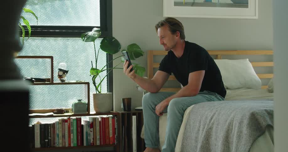 A man sits on his bed during a video call on his cellphone and shows the other person his living space