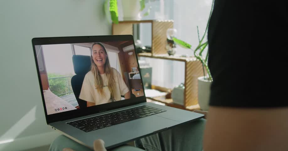 A man at home sits with a laptop on his knee and waves at the teenage waving to him on the screen
