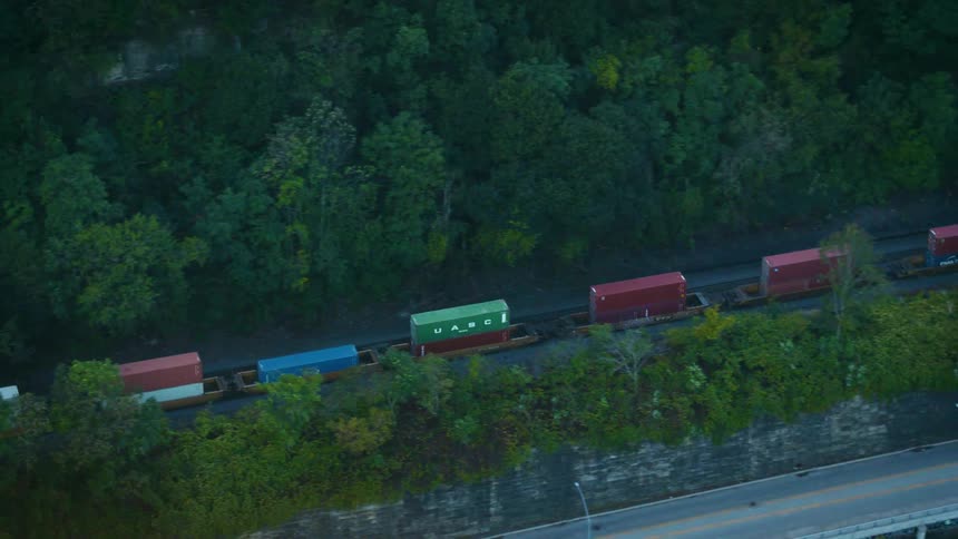 Train cars move through a forested area with a nearby highway