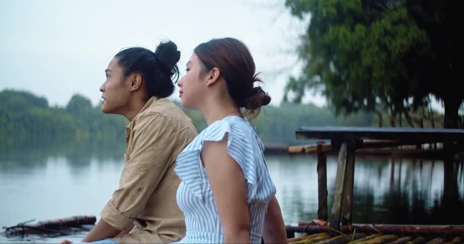 A man and a woman sitting by a lake with the trees the sky and clouds in the background