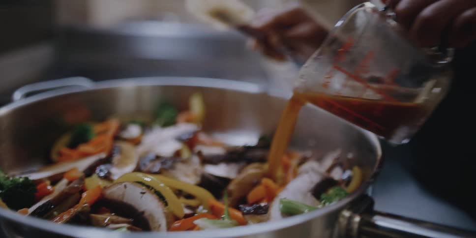 A female chef smiles as she pours sauce over the top of a mixture of ingredients in a heated pan