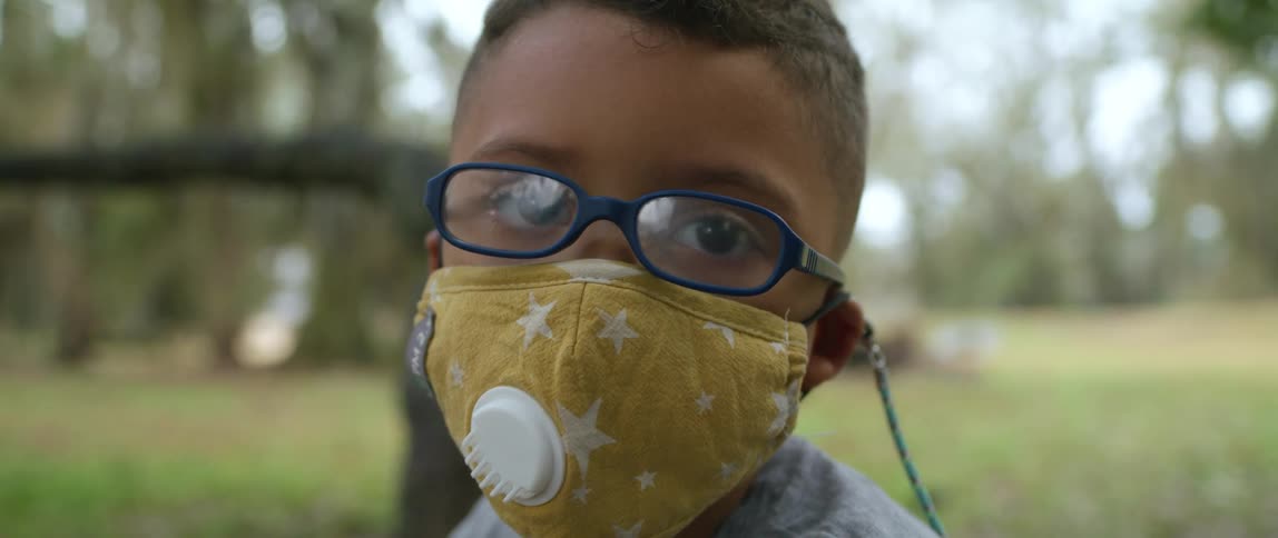 A young boy wearing glasses and a face mask looks directly towards the camera