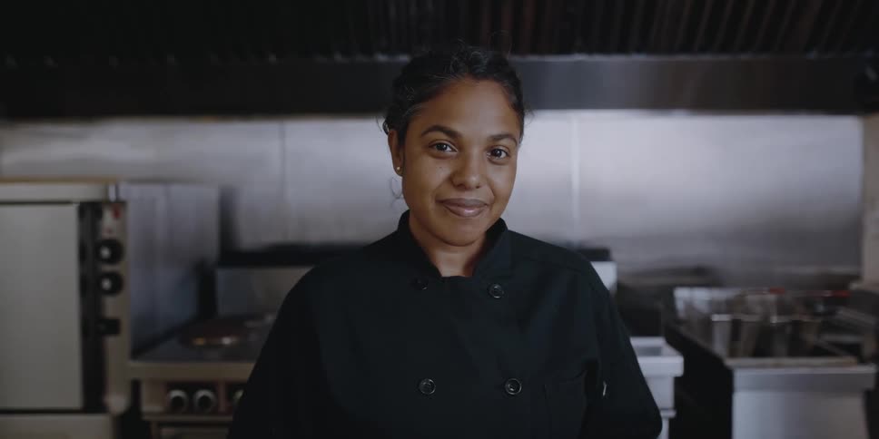 A women is wearing chef clothing in a kitchen while she looks at the camera and smiles