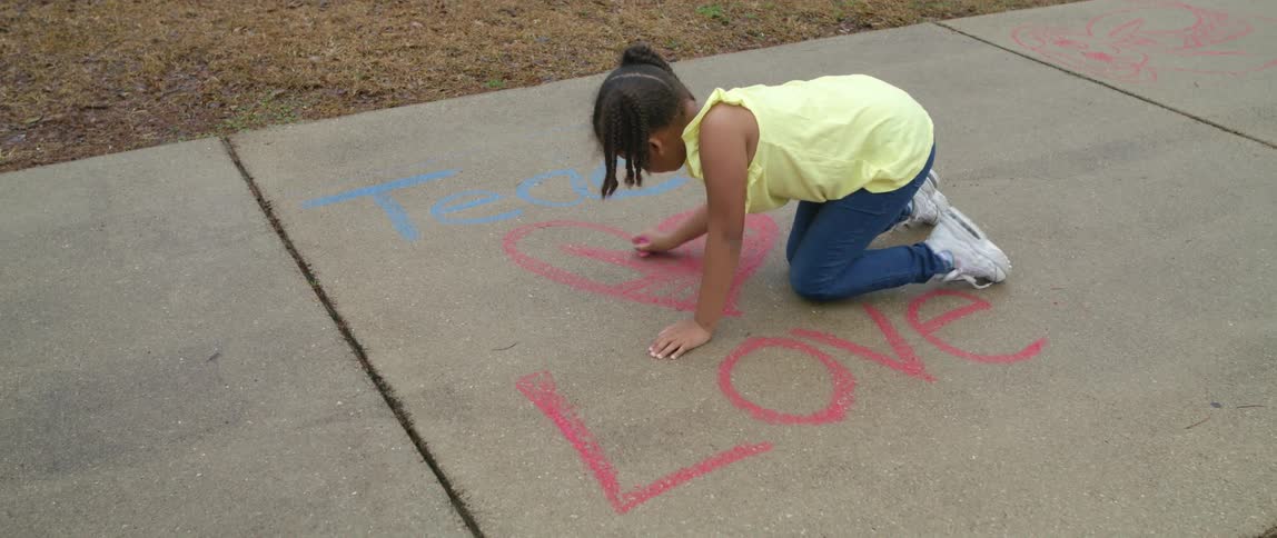 A young girl kneels on the ground coloring a sidewalk with chalk
