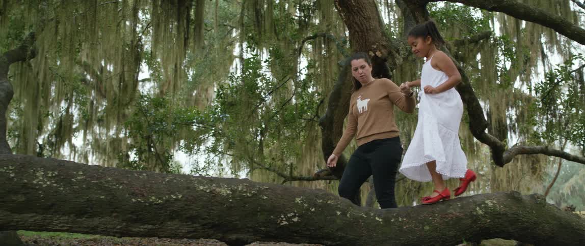 A woman holding a girls hand as she walks on a tree trunk with the sky and clouds in the background