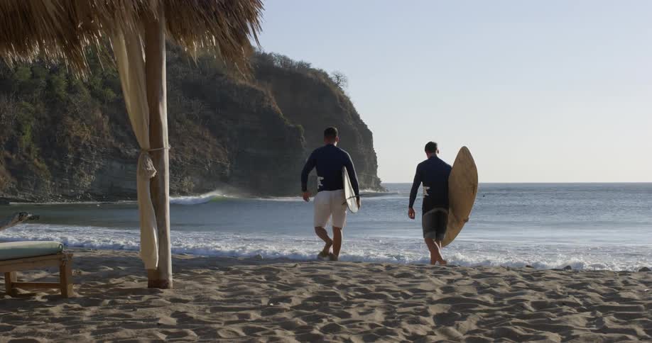 Two men walk across a beach carrying surfboards