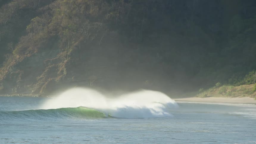 Large wave rolls in crashing to spray water into air near beach and forest covered mountain