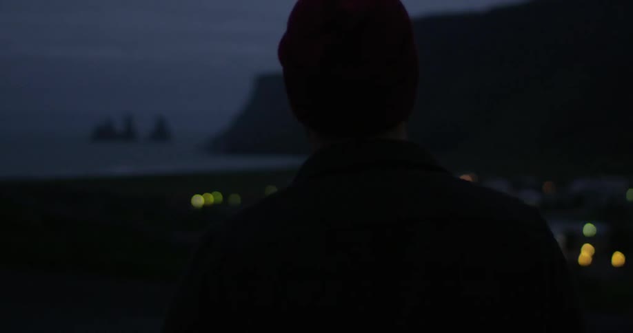 A man standing on a hilltop overlooking a small village and the coast at dusk