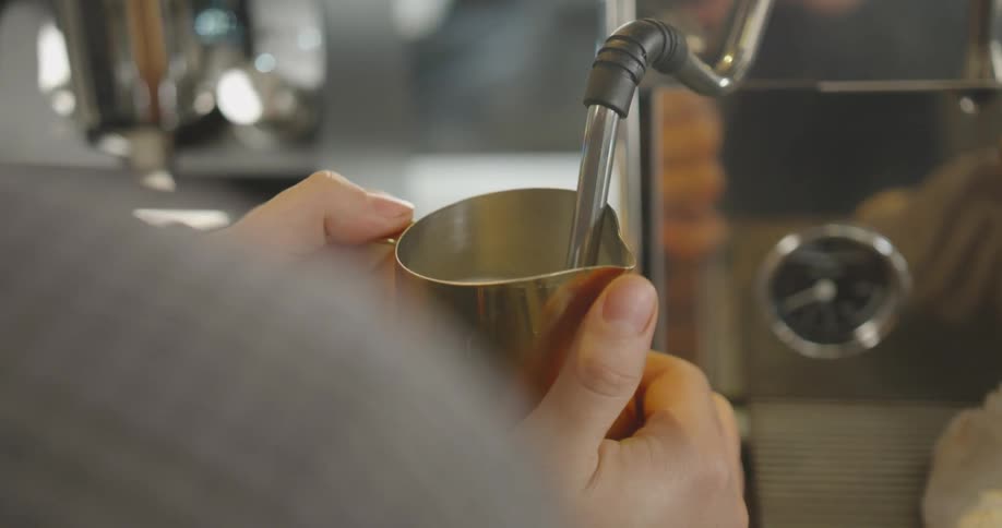 A barista froths milk in small metal pitcher in coffee shop