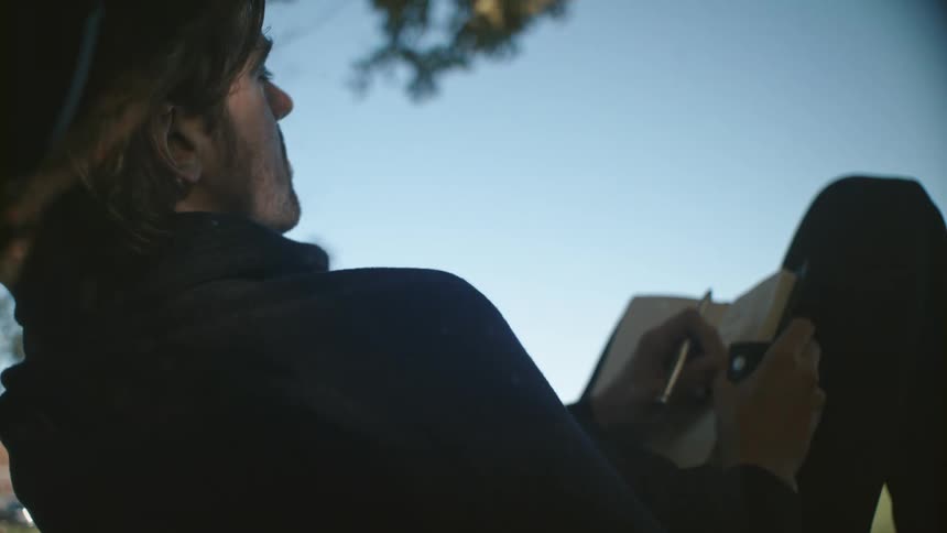 A man sitting by a tree with a pen and a book with the sky and clouds in the background