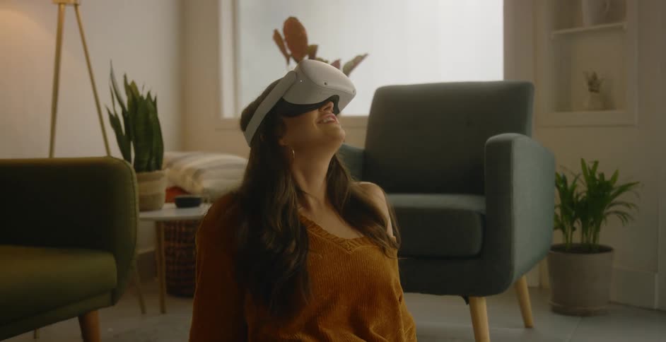 A young woman looks around while wearing a virtual reality headset while sitting on the carpet of the living room of a house during the day