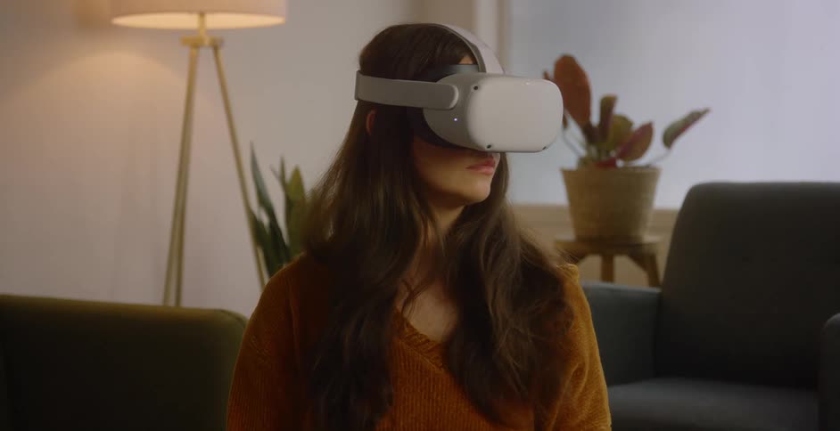 A woman sits in the living room of a house with a virtual reality headset on her face