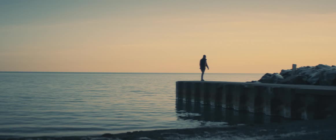 A silhouette of a person walking on a pier with a body of water the sky and clouds in the background