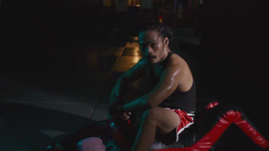 Sweat covered boxer sits on cushion near boxing gloves in shadowed room