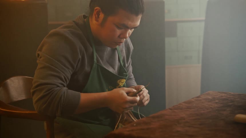 A shoe maker sits in a back room lined with shoe boxes and uses a leathering tool and thread