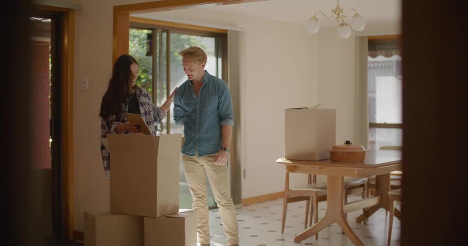 A man carries a box in to a house and sets it on a table and a woman shows him a photo she pulls out of a box and they both laugh