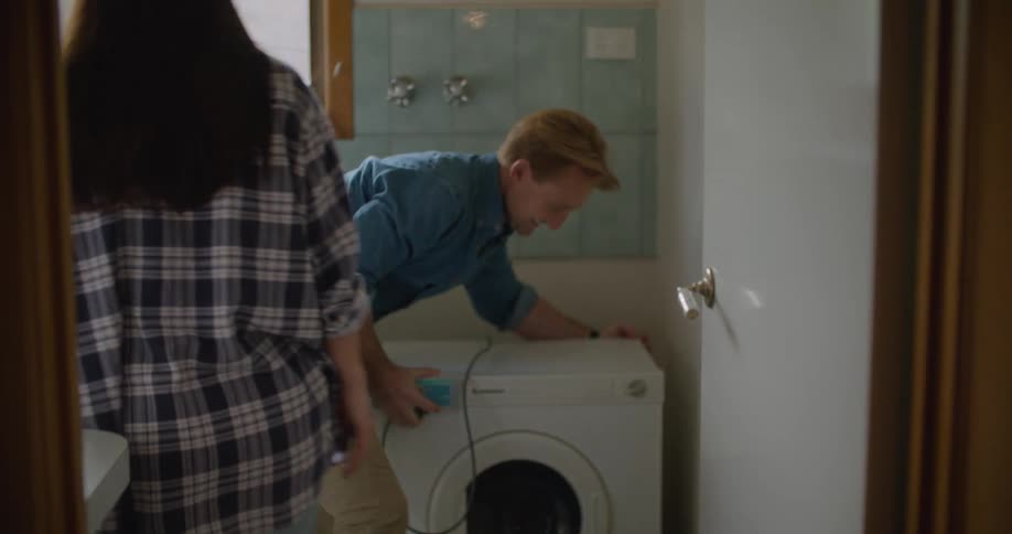 A young woman adjusts a curtain on a large window before walking into another room where a man is moving and plugging in a washing machine